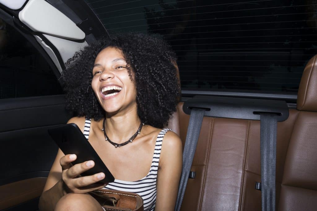 Portrait of laughing woman with smartphone sitting on the rear seats of a car